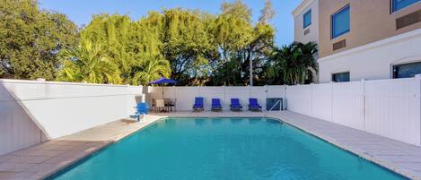 A swimming pool with blue lounge chairs, a sun umbrella, and a white fence.
