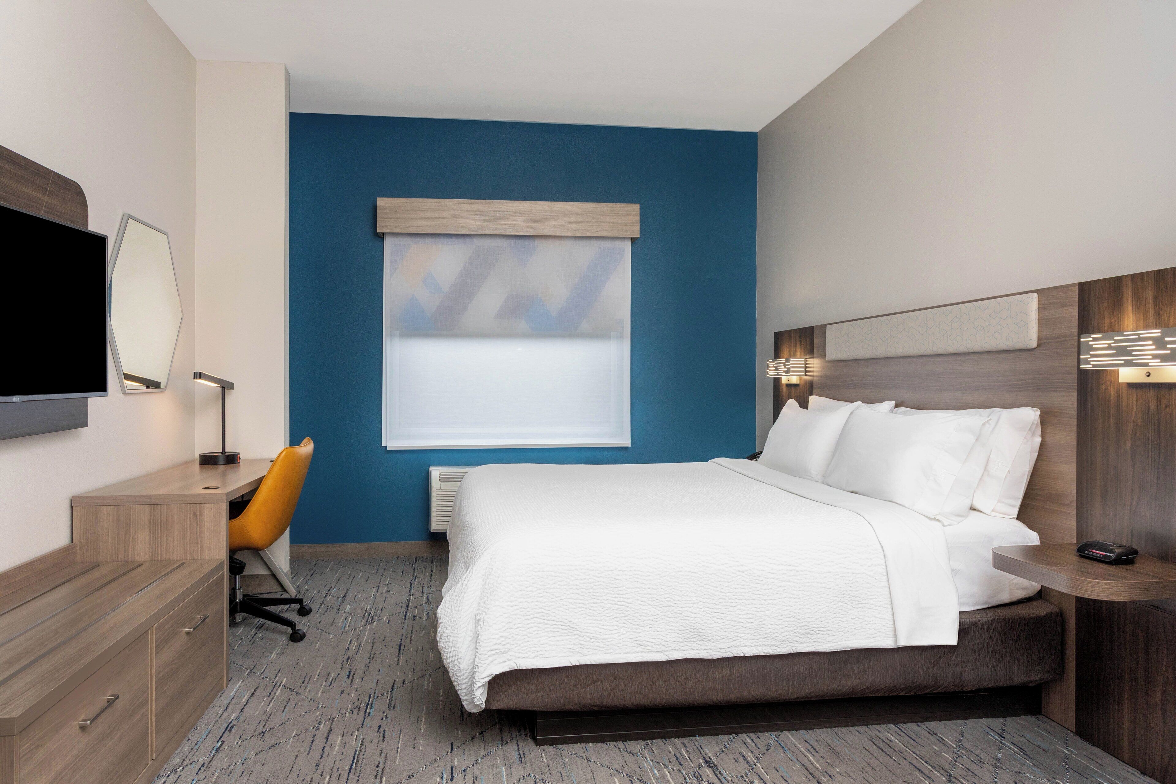 A hotel room with a bed, a desk with a chair, a TV, and a window with blinds.