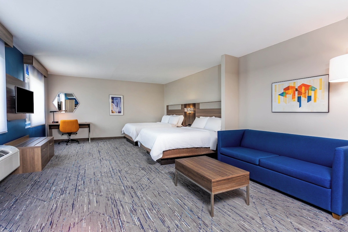 A hotel room with a bed, a blue sofa, a desk with a chair, a TV, and artwork on the walls.