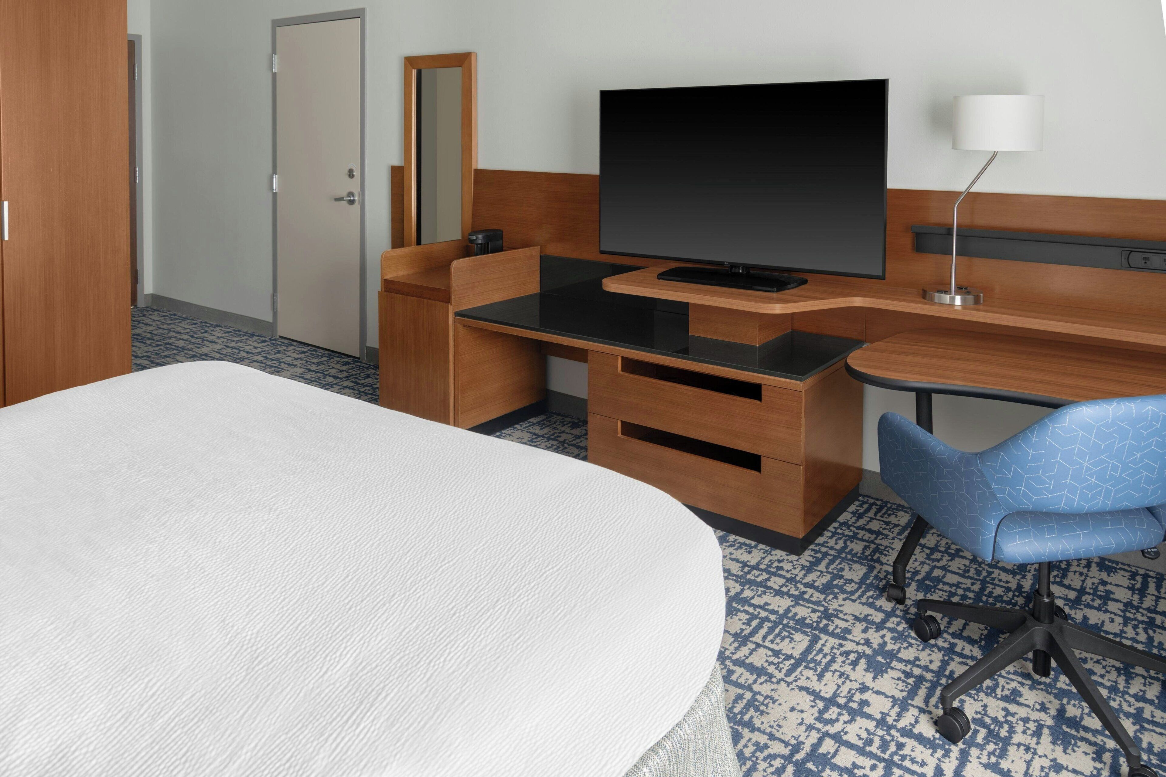 Quarto de hotel com cama, escrivaninha com televisão de tela plana, cadeira, abajur e cômoda.