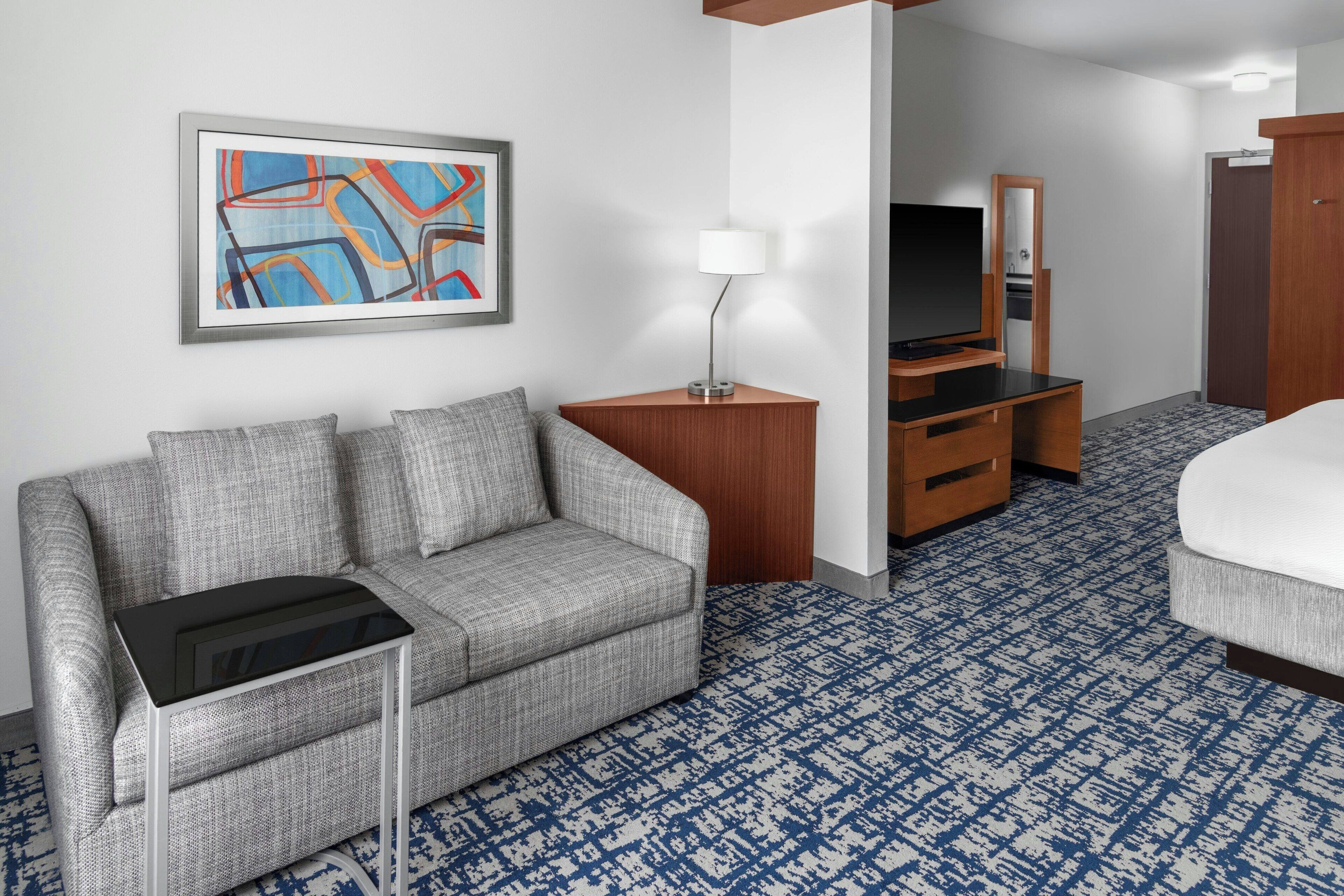 Habitación de hotel con sofá gris, una mesita pequeña, un televisor sobre un mueble de madera, un espejo y una cama con colchón blanco.