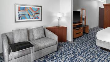 Habitación de hotel con sofá gris, una mesita pequeña, un televisor sobre un mueble de madera, un espejo y una cama con colchón blanco.