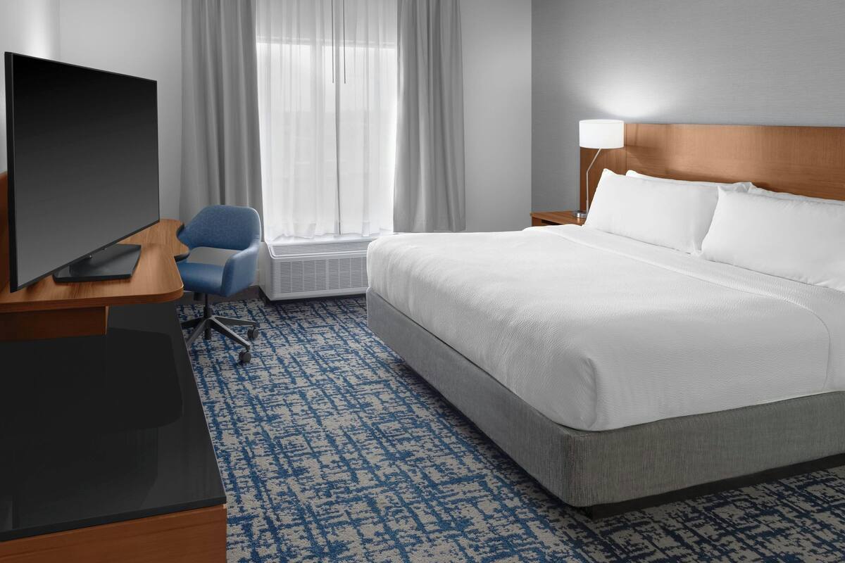 A hotel room with a large bed, a desk with a computer, a chair, a TV, and a window with curtains.