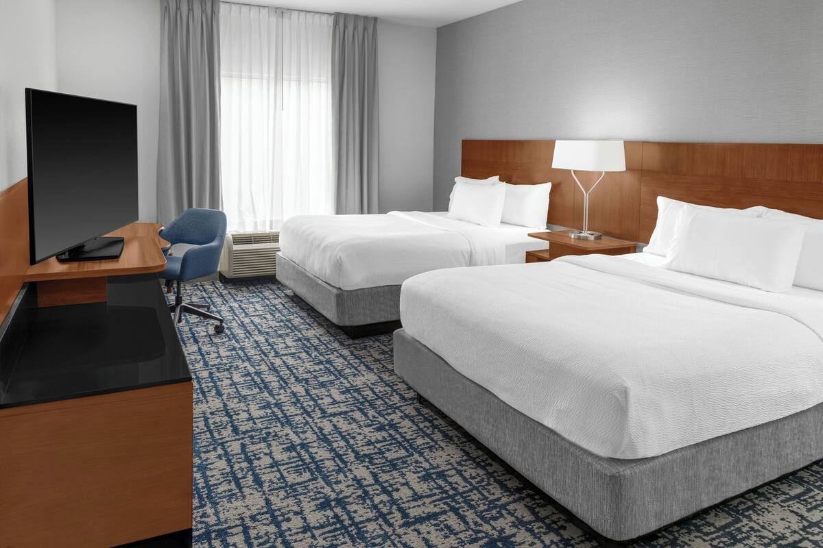 A hotel room with two beds, a desk with a flat-screen TV, a chair, a lamp, and a large window with curtains.