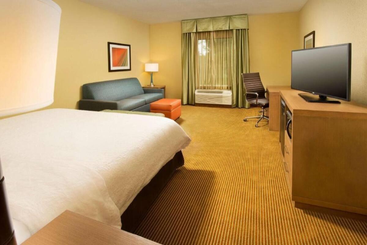 Ein Hotelzimmer mit einem Bett, einer Couch, einem Fernseher, einem Schreibtisch und einem Vorhangfenster.
