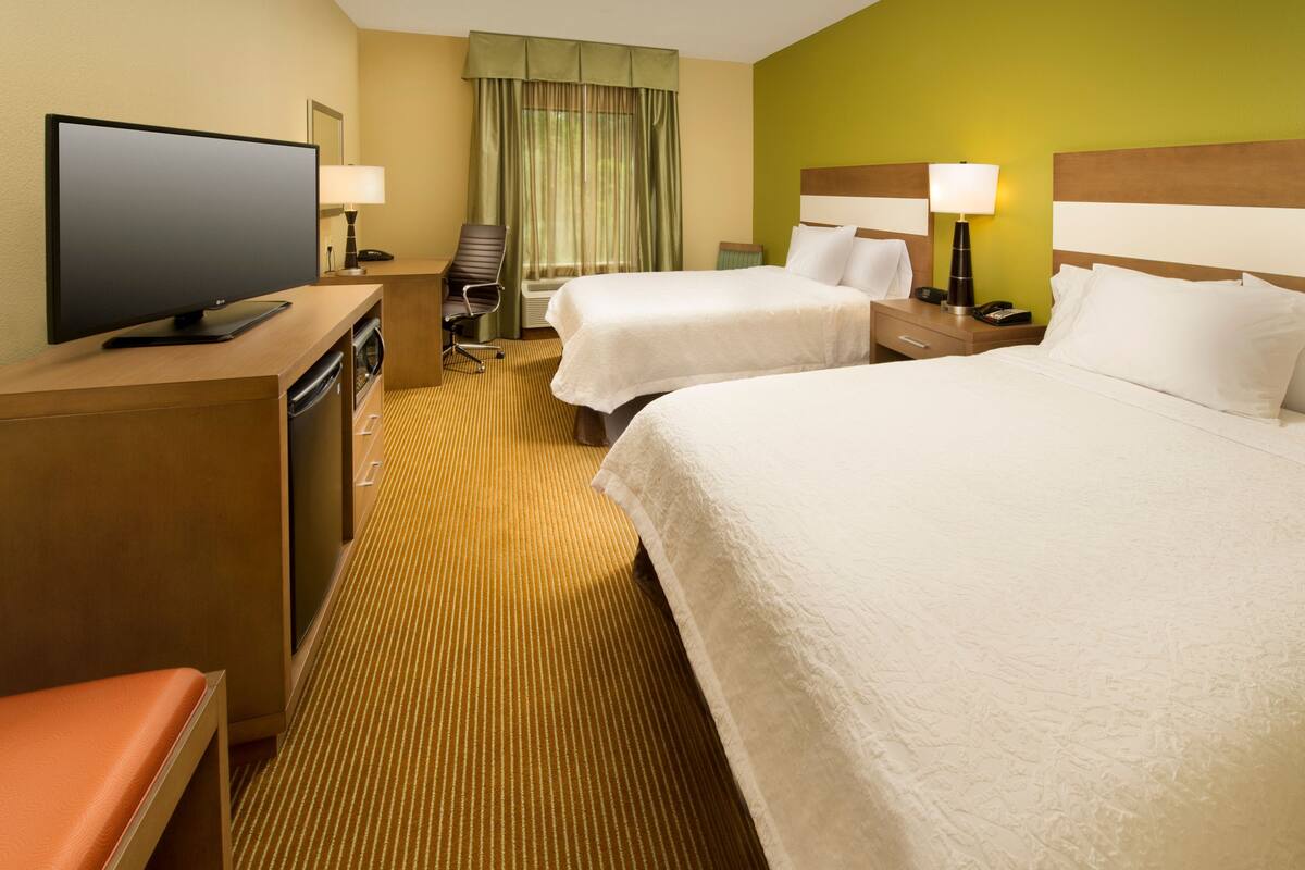 Ein Hotelzimmer mit zwei Betten, einem Fernseher, einem Schreibtisch mit Stuhl und einem Fenster mit Vorhängen.