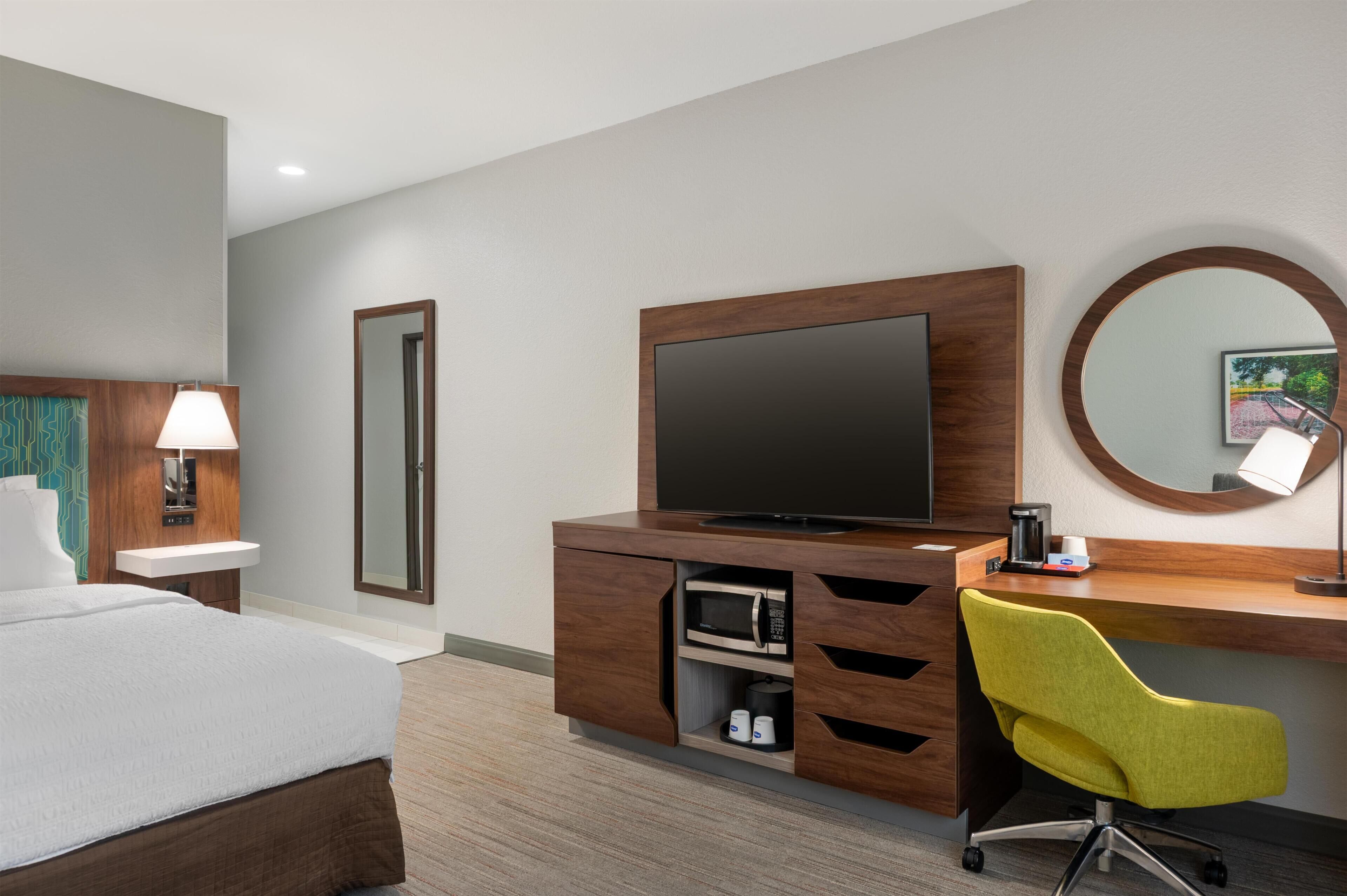 Habitación de hotel con cama, escritorio con silla, televisor, espejo, lavamanos y microondas.