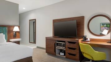 Habitación de hotel con cama, escritorio con silla, televisor, espejo, lavamanos y microondas.