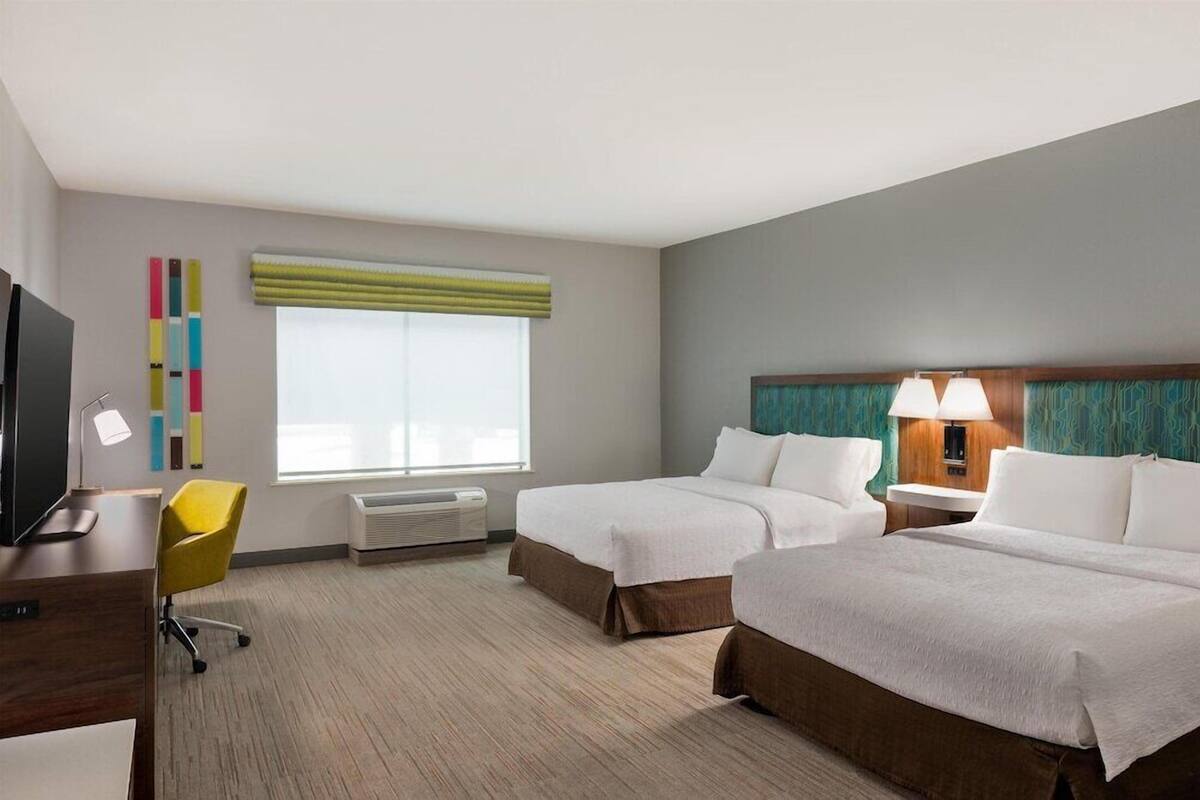 Ein Hotelzimmer mit zwei Betten, einem Schreibtisch, einem Stuhl, einem Fernseher, einem Fenster mit Jalousien, einer Klimaanlage und einer Lampe.