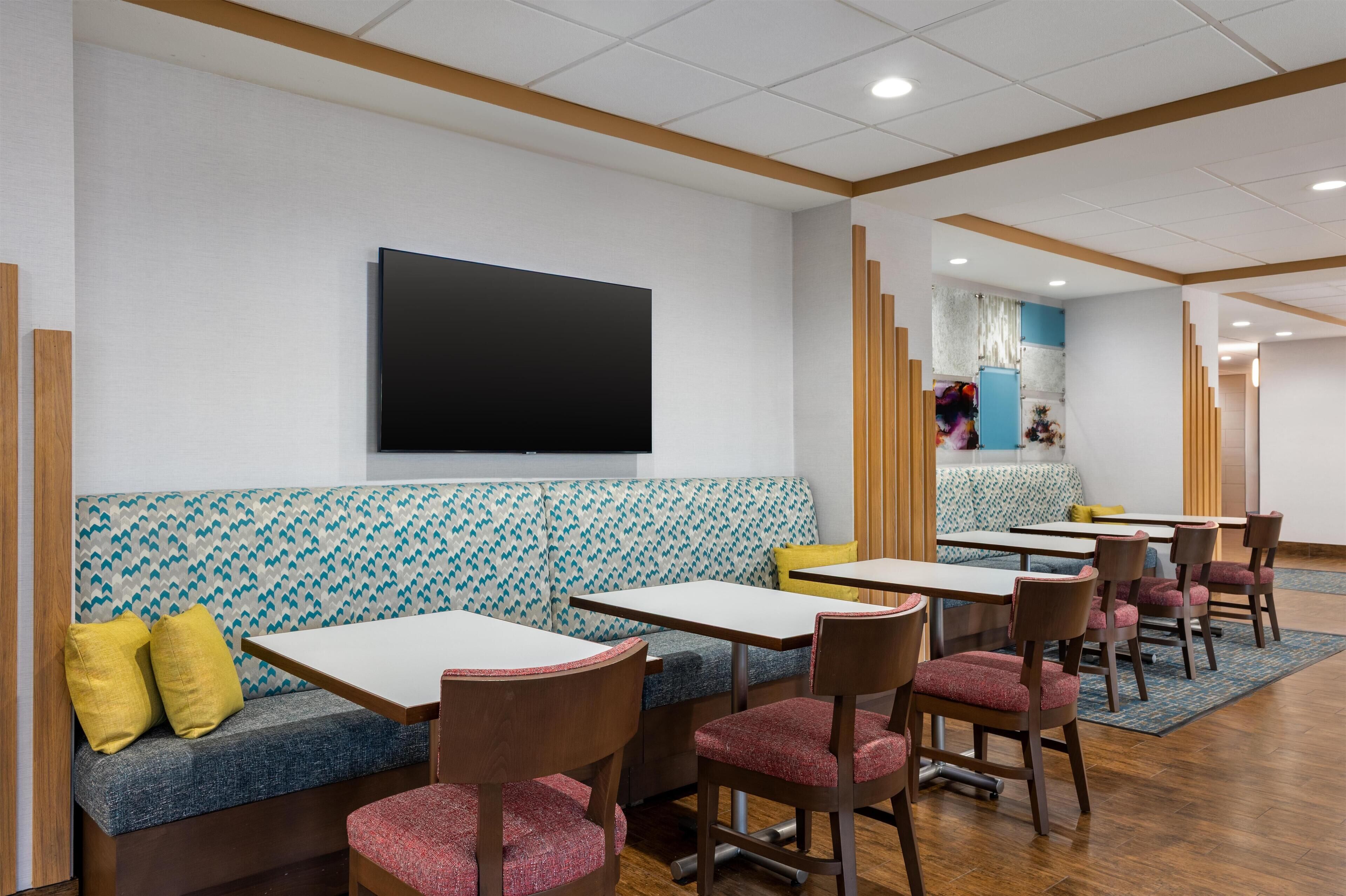A modern interior with a large flat-screen TV mounted on the wall, a patterned sofa with yellow cushions, and several tables and chairs arranged for dining.