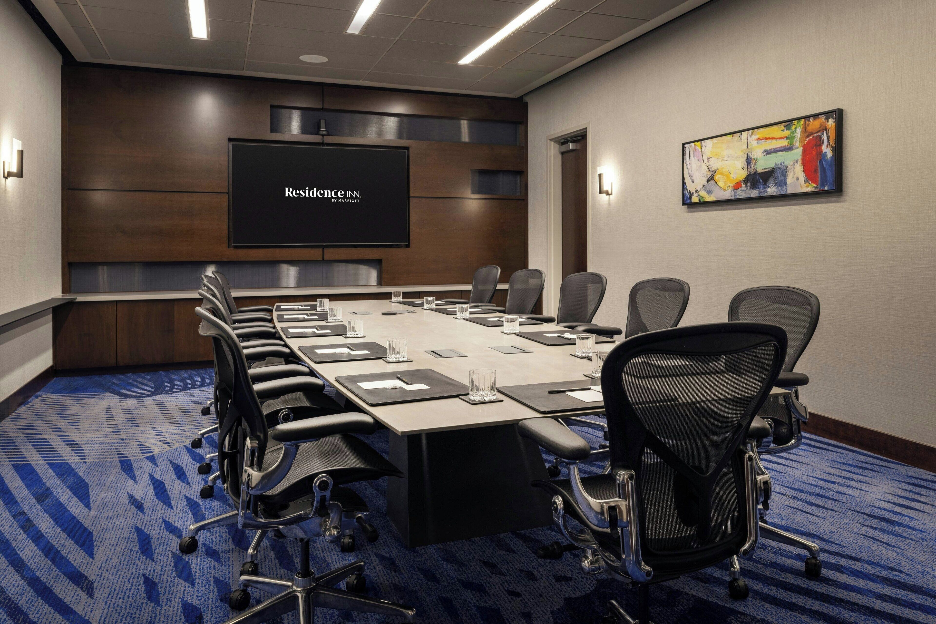 A conference room with a long table, multiple chairs, a large screen displaying "Residence Inn," and a colorful abstract painting on the wall.