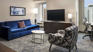 Habitación de hotel con un sofá azul, un sillón estampado, un televisor de pantalla plana, una mesita redonda y vista a un paisaje urbano a través de un ventanal grande.