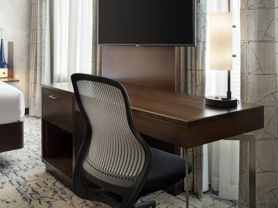 Une chambre d’hôtel avec un bureau en bois, une chaise moderne, un téléviseur à écran plat, une table de chevet avec une lampe et une moquette à motifs.