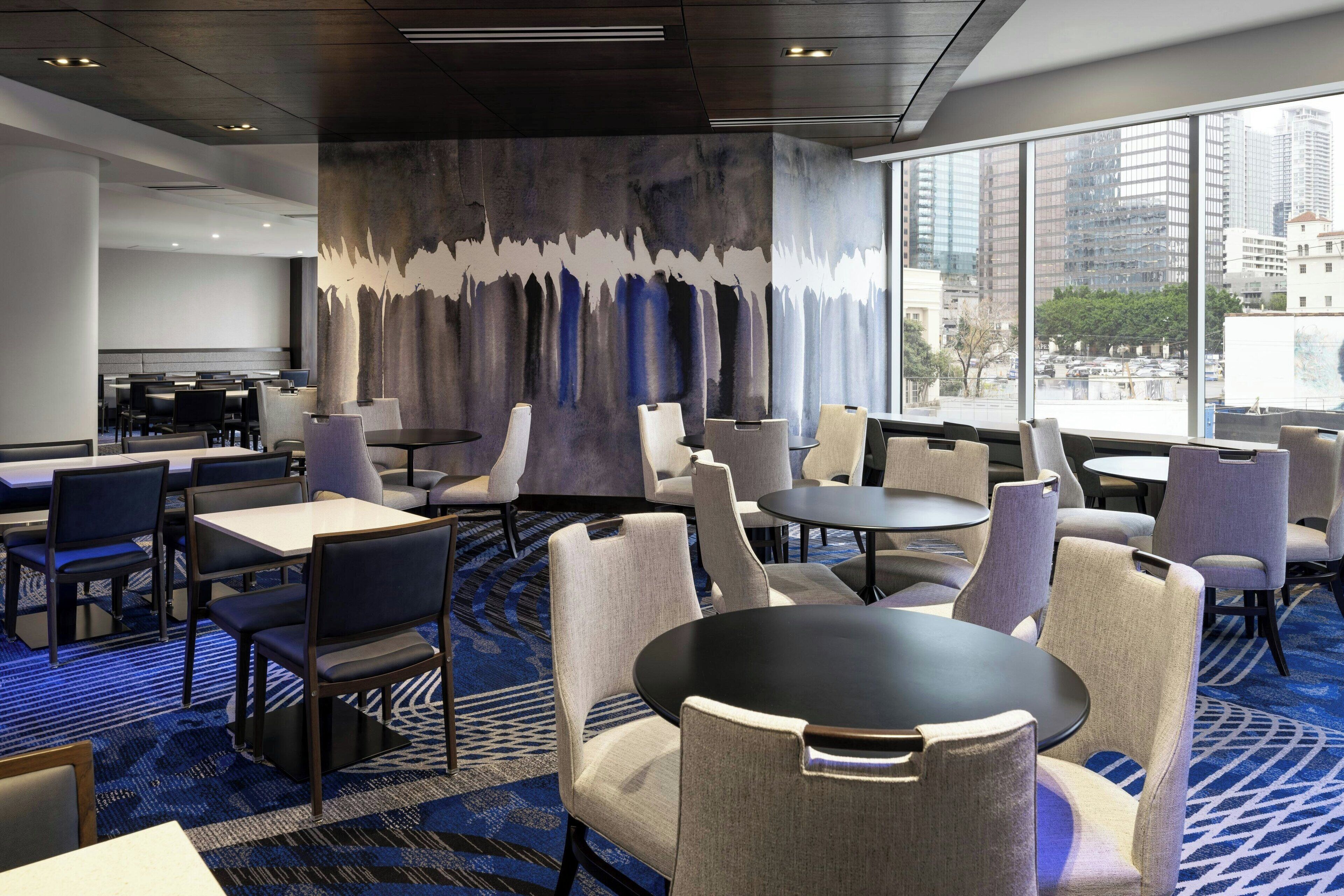 Un vestíbulo de hotel con diseño moderno, que presenta una variedad de disposiciones de asientos, incluyendo mesas y sillas, un gran mural abstracto en la pared y amplias ventanas con vista a la ciudad.