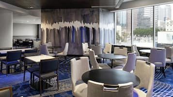 Un vestíbulo de hotel con diseño moderno, que presenta una variedad de disposiciones de asientos, incluyendo mesas y sillas, un gran mural abstracto en la pared y amplias ventanas con vista a la ciudad.
