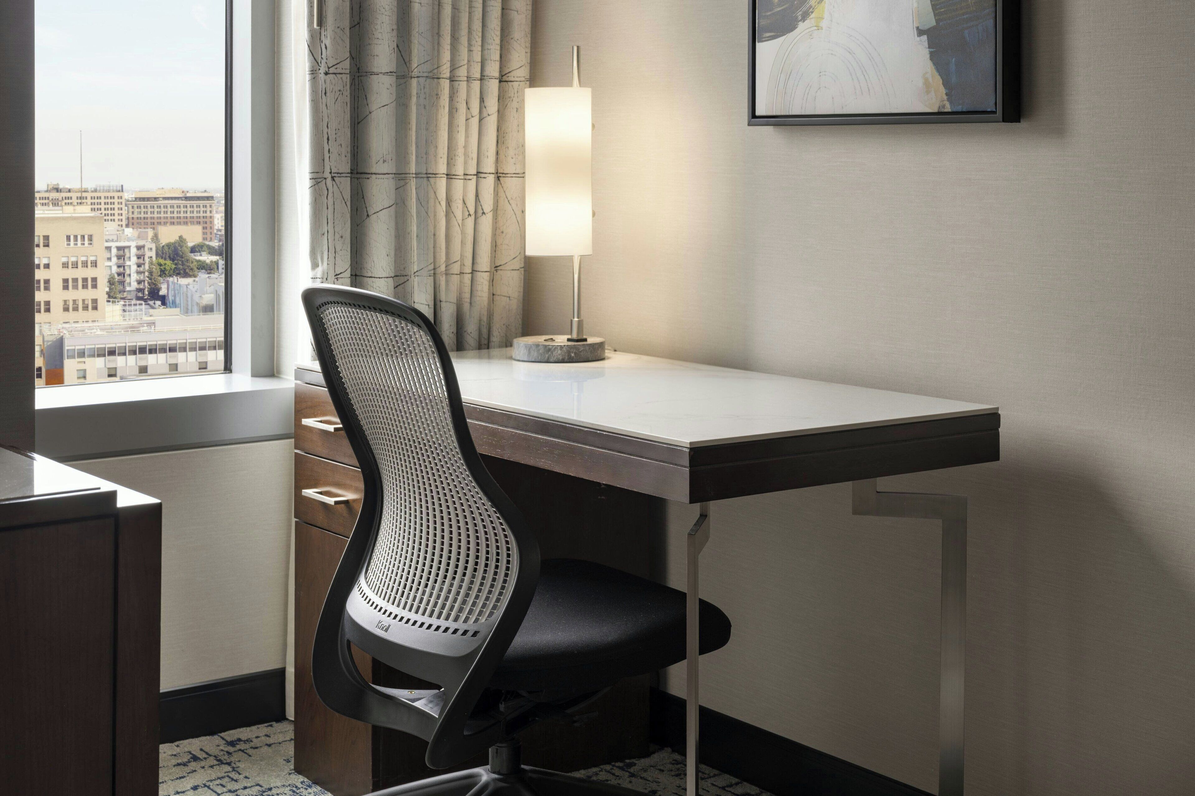 Hotelkamer met een bureau, stoel, lamp, raam met uitzicht op gebouwen, en een lijst aan de muur.