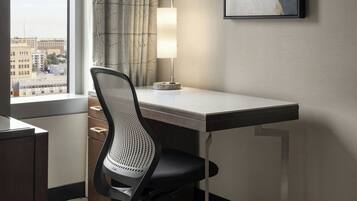 Hotelkamer met een bureau, stoel, lamp, raam met uitzicht op gebouwen, en een lijst aan de muur.
