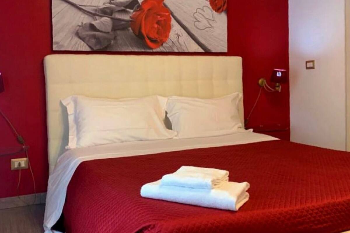 A hotel room with a red wall, a bed with white bedding, a red blanket, and a heart-shaped ottoman.