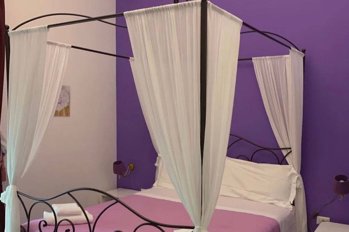 A four-poster bed with white drapes, a purple wall, and a wooden floor.