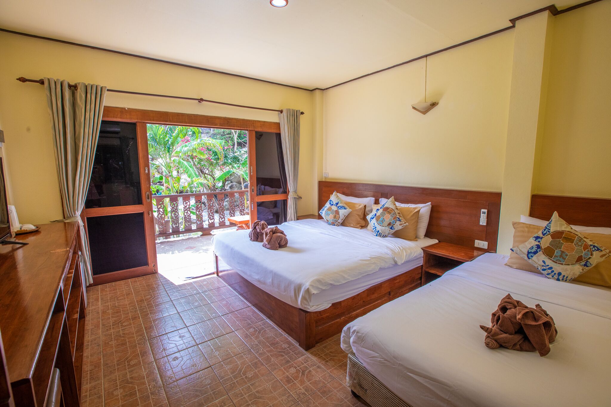 A hotel room with two beds, a wooden desk, a balcony with curtains, and a view of greenery.