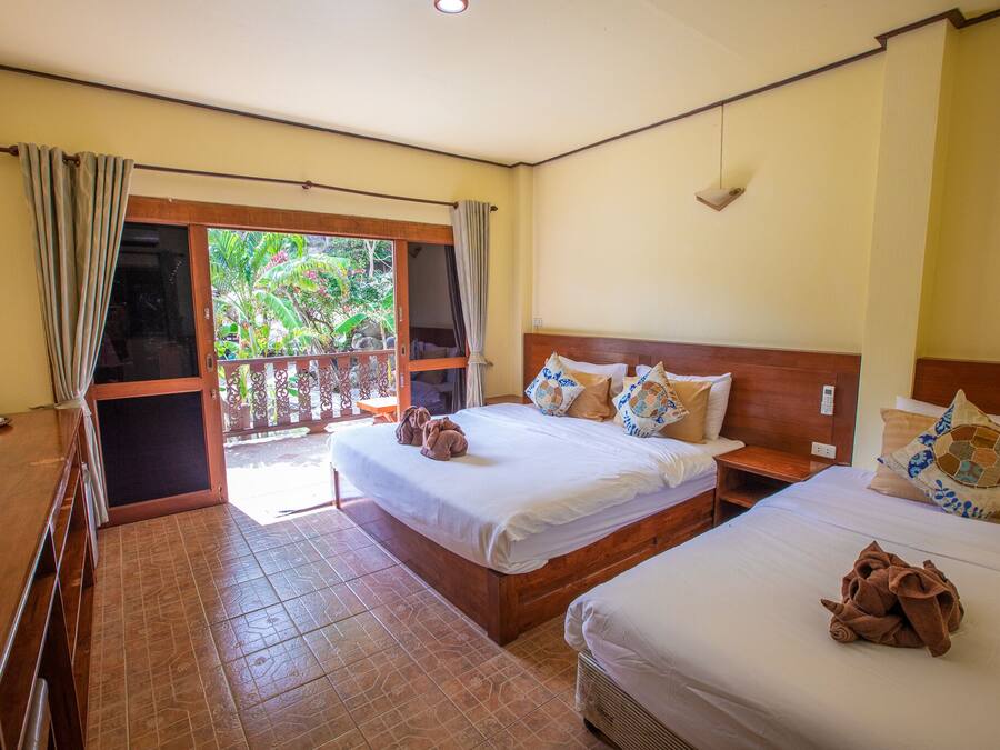 A hotel room with two beds, a wooden desk, a balcony with curtains, and a view of greenery.
