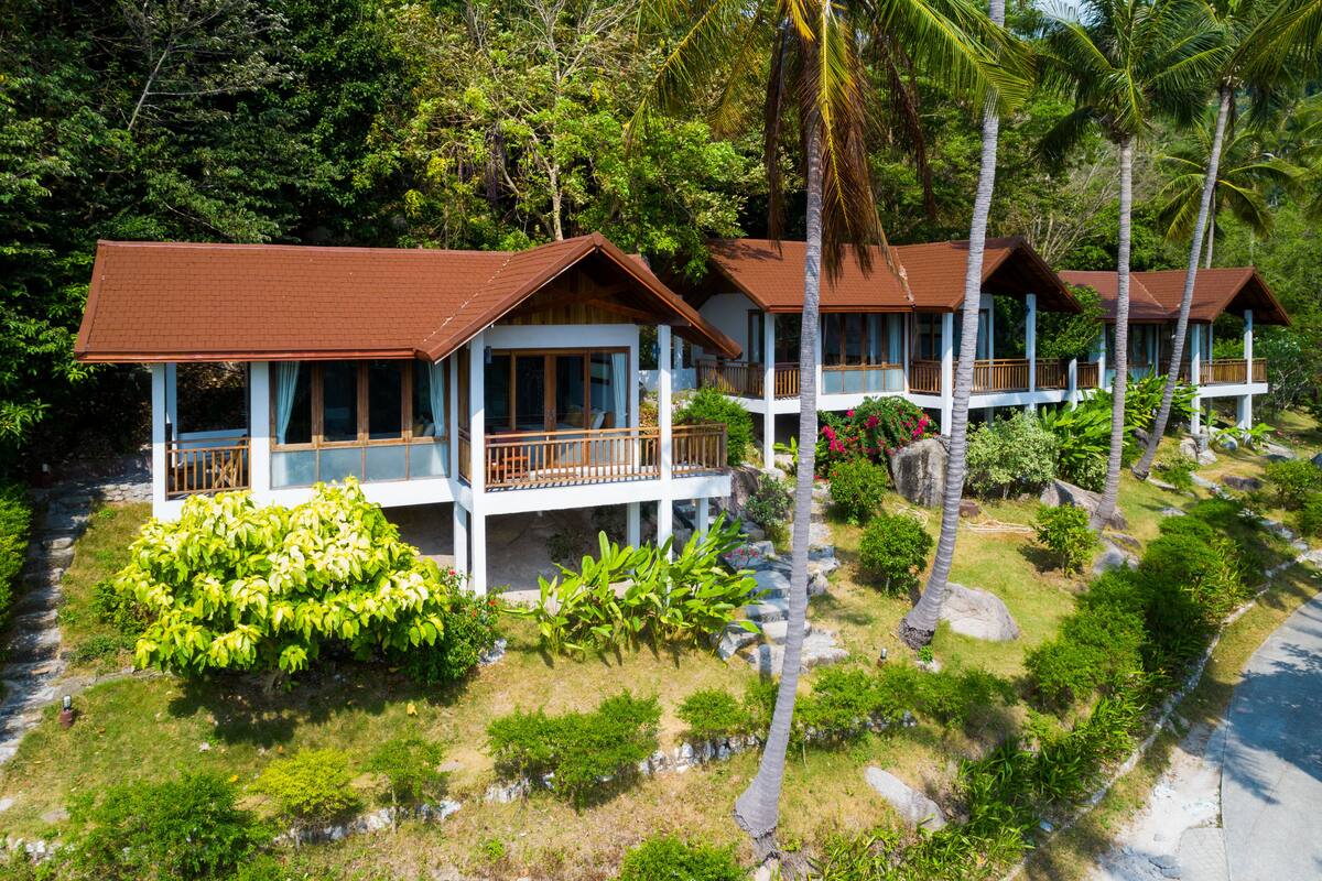 A row of two-story beachfront cottages with balconies, surrounded by lush greenery and palm trees.