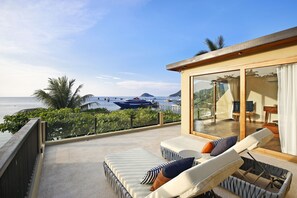 Un balcón con sillones de descanso y una mesa con vista al mar, con embarcaciones y vegetación al fondo.