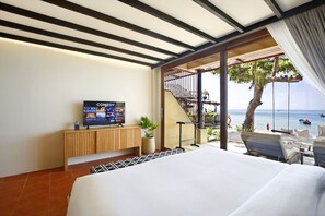 Habitación de hotel con cama, televisor sobre una consola de madera, balcón con vistas al mar y mobiliario exterior.