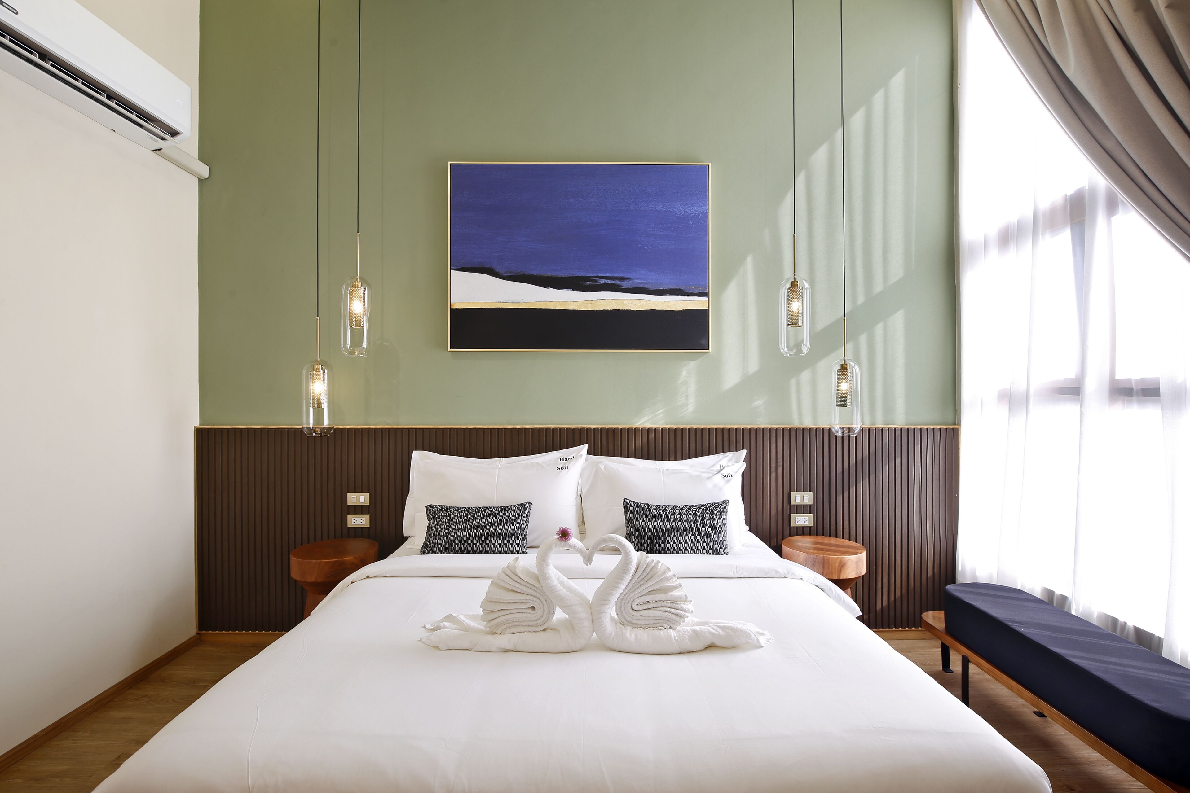 A hotel room with a bed, two bedside tables, a round wooden table, a wall-mounted painting, and hanging pendant lights.