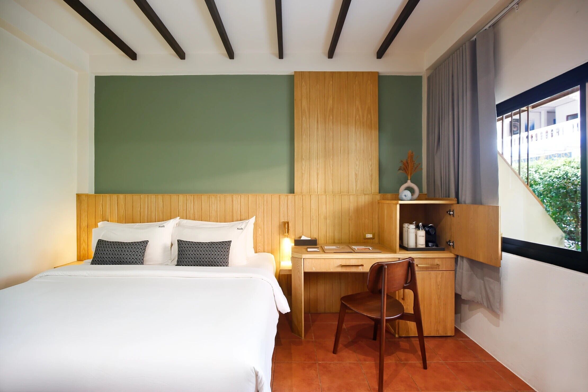 Habitación de hotel con cama, escritorio y silla. La pared es verde, el cabecero de madera y tiene una ventana con cortinas.