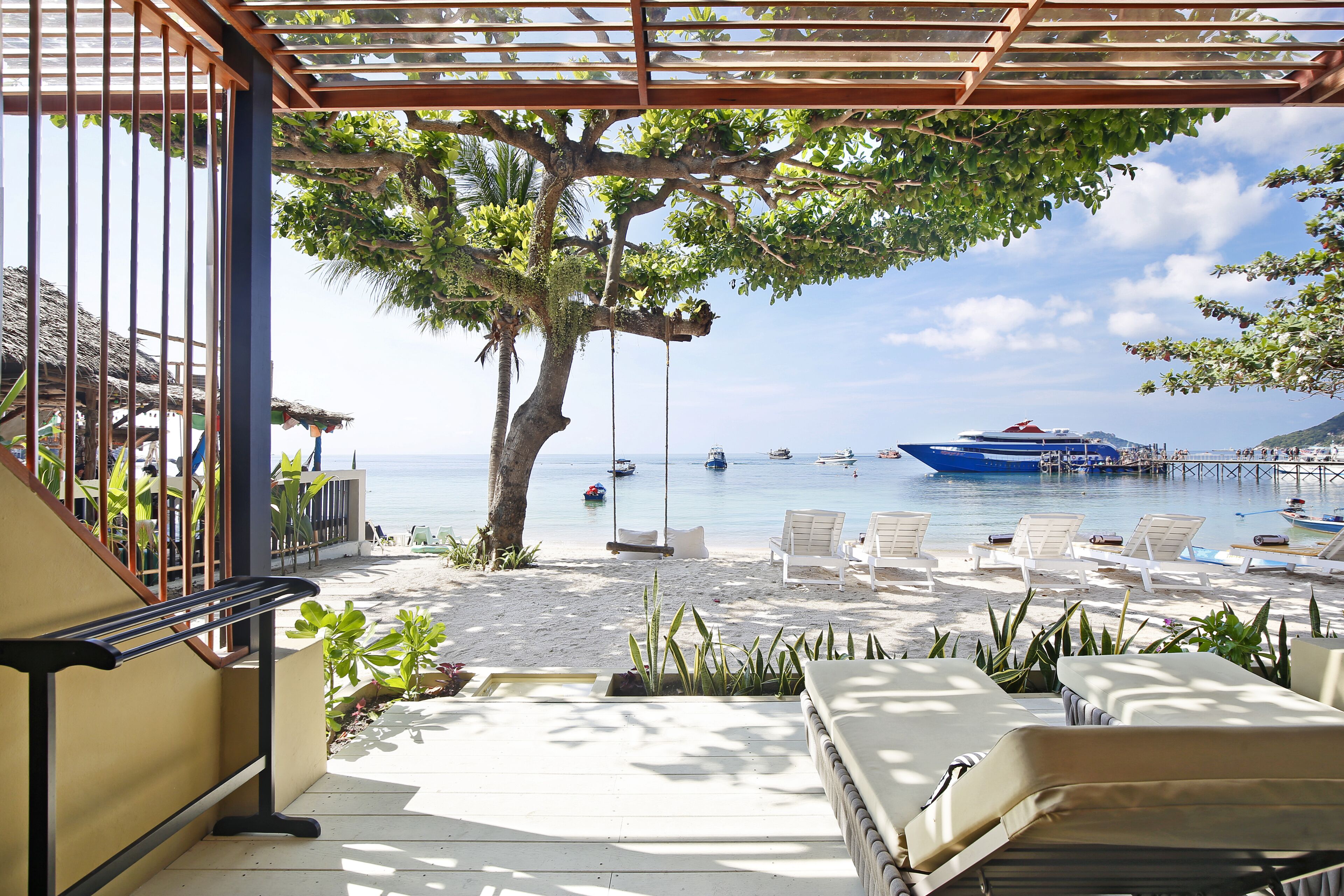 A beachside patio with lounge chairs, a swing, and a view of the sea and boats.