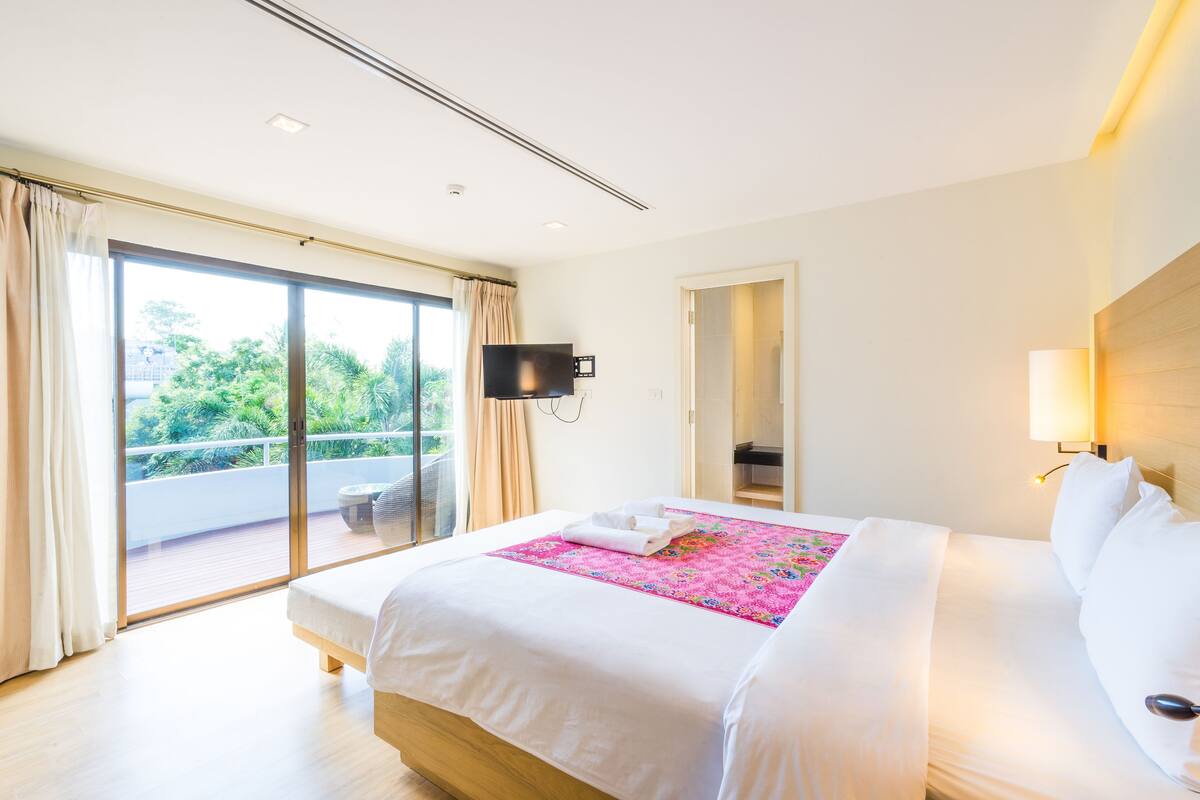 A hotel room with a large bed, a TV mounted on the wall, and a balcony with a view of greenery.