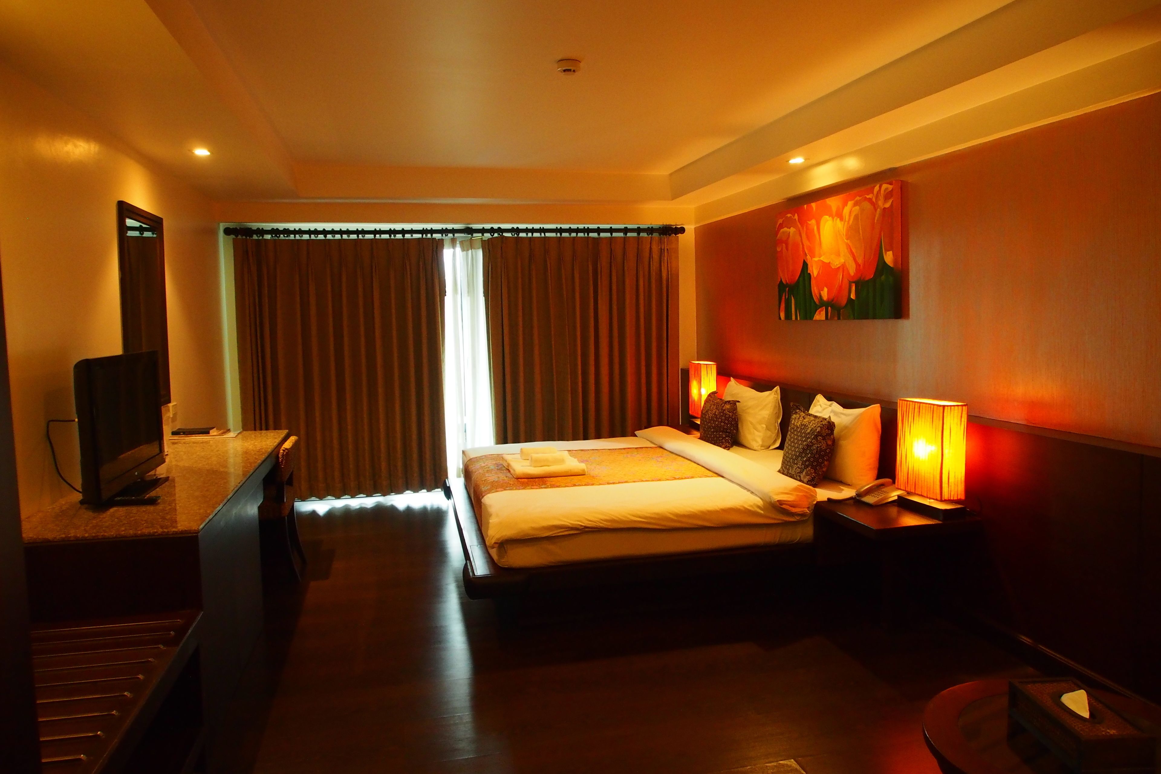 A hotel room with a bed, a TV, a desk, and a painting on the wall.