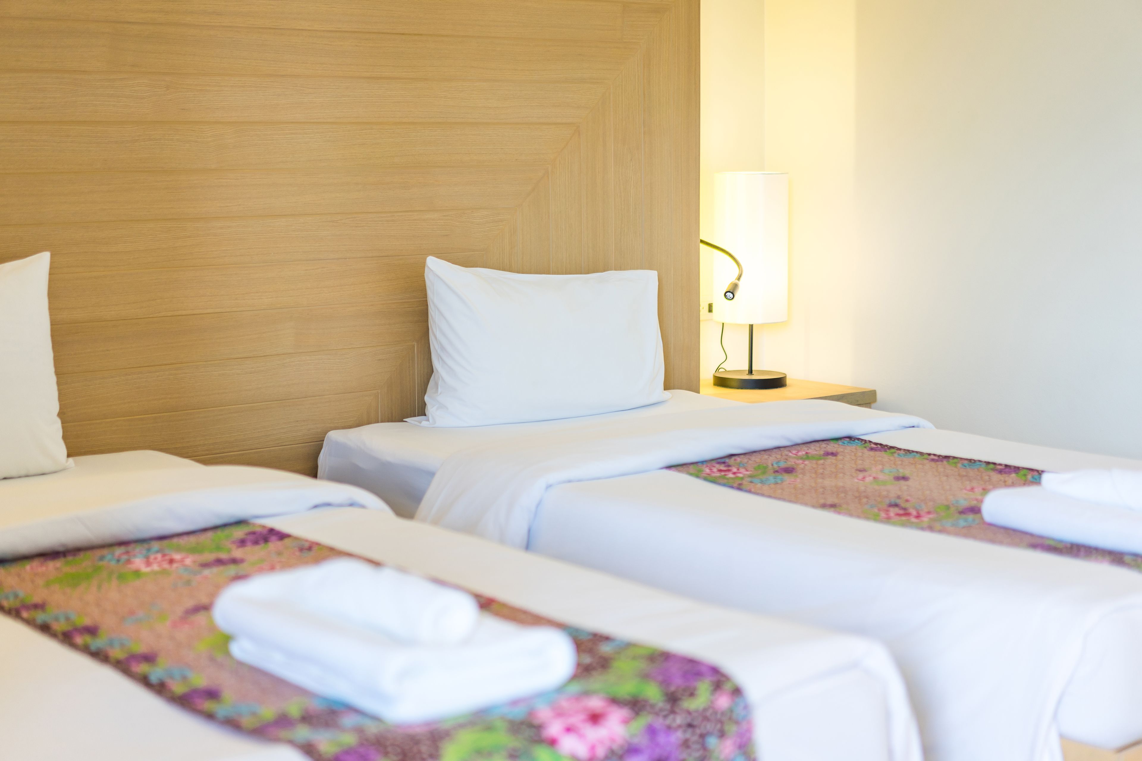 A hotel room with two beds, each with white linens and floral-patterned bedspreads. There is a nightstand with a lamp between the beds.