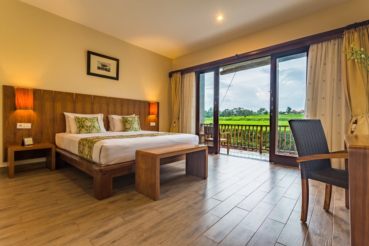 A bedroom with a wooden headboard, a bed with white linens, a bench, a chair, a nightstand, and a large sliding glass door leading to a balcony with a view of green fields.