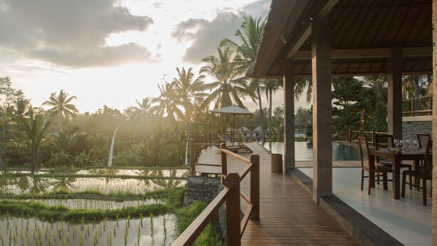 A wooden deck with a view of a rice field and palm trees, a covered dining area with tables and chairs, and a stone structure in the background. - Manyi Village (Ubud)