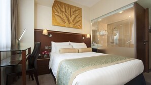 A hotel room with a large bed, a desk with a lamp, a chair, a nightstand with a vase of flowers, a large mirror, and a shower area. - Brighton Hotel Hong Kong (Hong Kong)