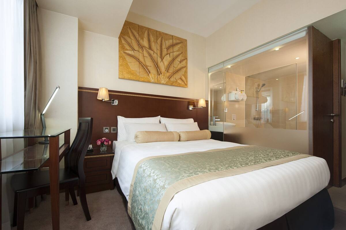 A hotel room with a large bed, a desk with a lamp, a chair, a nightstand with a vase of flowers, a large mirror, and a shower area.