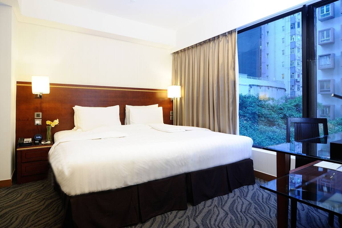 A hotel room with a large bed, two bedside lamps, a nightstand with a phone and a vase of flowers, a glass table, and a view of a cityscape through a large window.