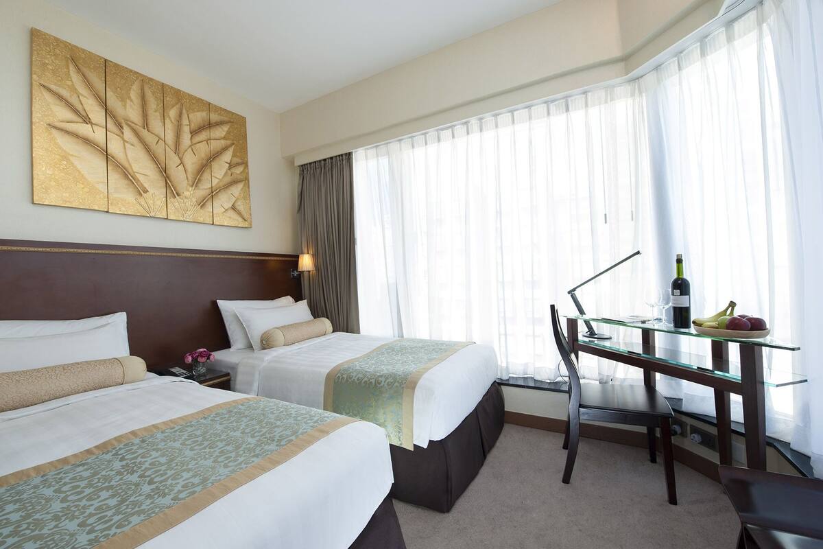 A hotel room with two beds, a glass table with a bottle and fruit, and a large window with sheer curtains.