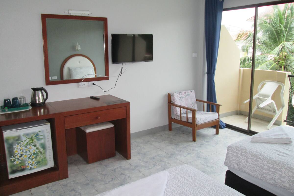 A hotel room with a wooden desk, a chair, a bed, a TV, and a balcony with a view of palm trees.