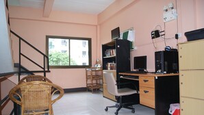 Una habitación con escritorio, computadora, estantería, silla de mimbre y ventana con vista a edificios.