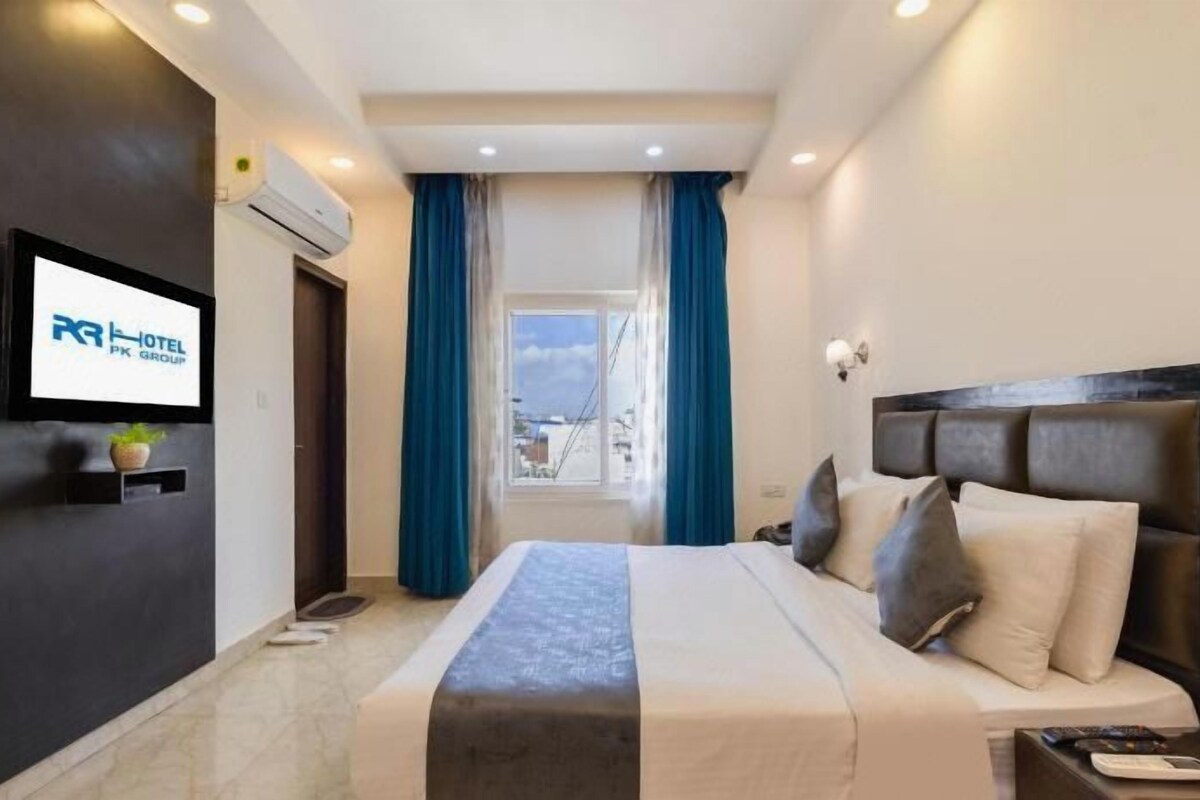A hotel room with a bed, a TV mounted on the wall, a window with curtains, and a view of the outdoors.