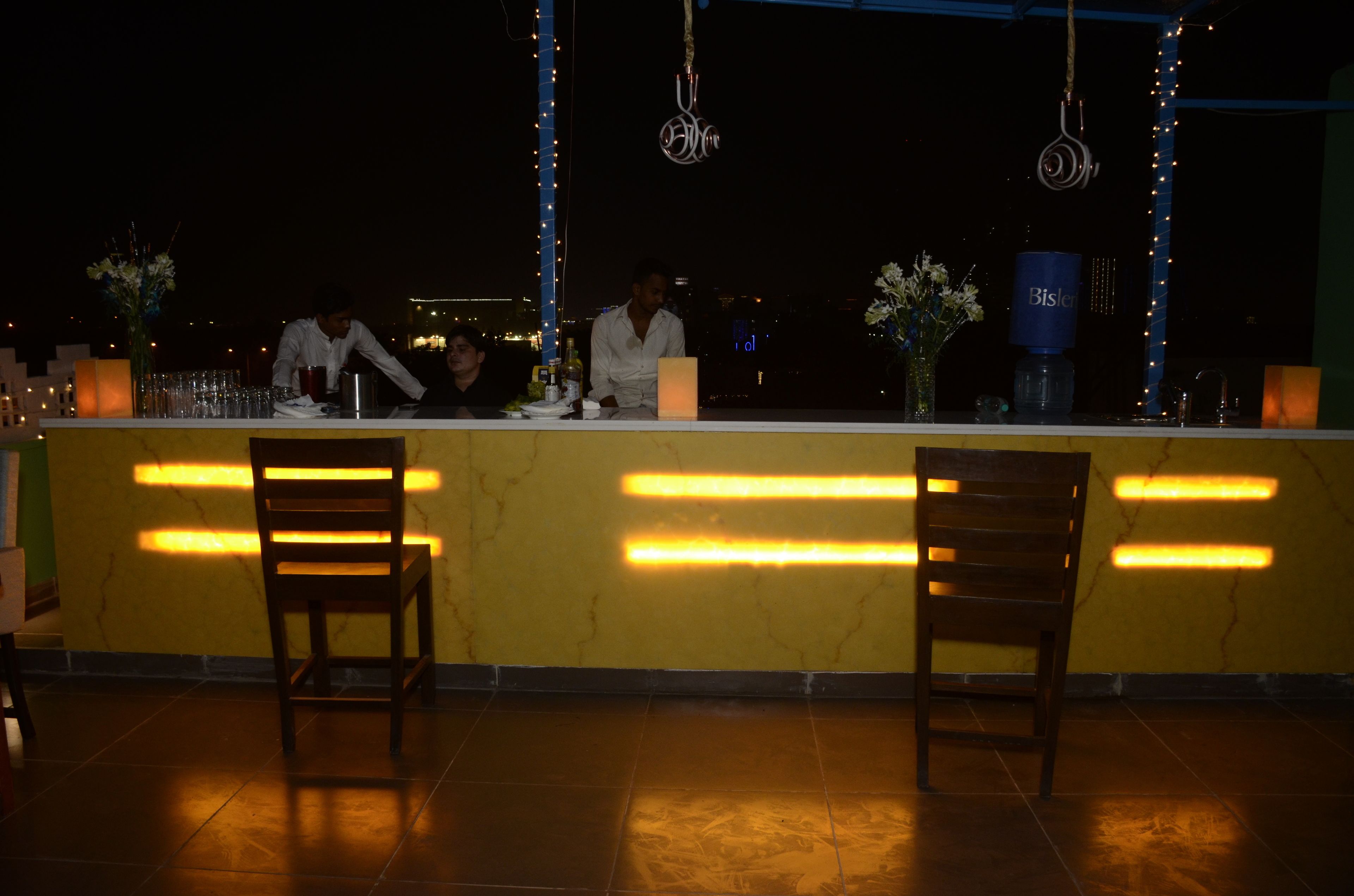 A bar area with a yellow counter, two wooden chairs, and a city view at night.