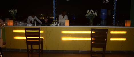 A bar area with a yellow counter, two wooden chairs, and a city view at night.