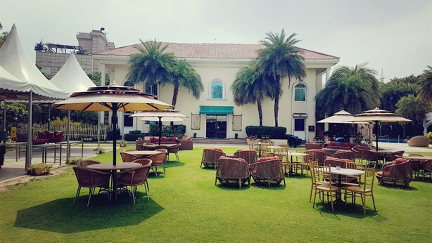An outdoor seating area with tables and chairs, surrounded by palm trees and a building with a tiled roof. - The Palms - Town & Country Club (Gurugram)