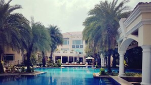 A large indoor swimming pool surrounded by palm trees and a building with multiple windows. - The Palms - Town & Country Club (Gurugram)