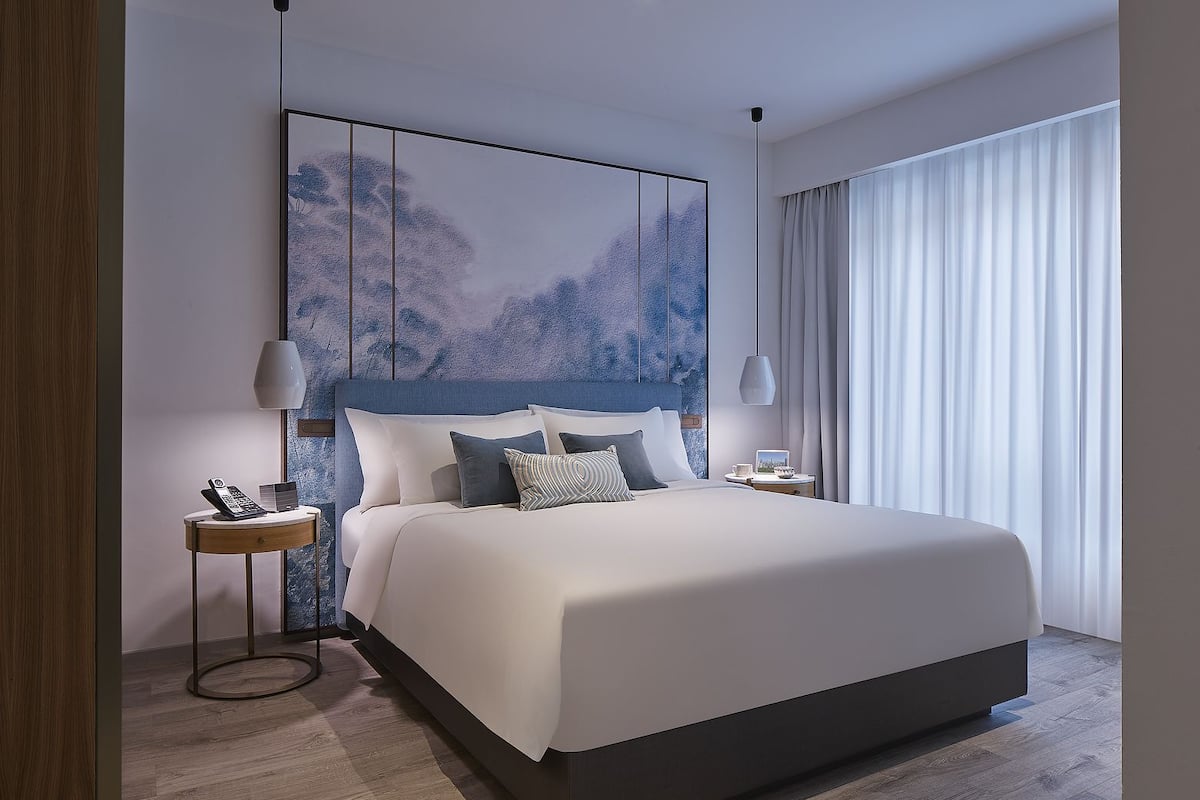 A modern hotel room with a large bed, a bedside table with a phone, a lamp, and a painting on the wall.