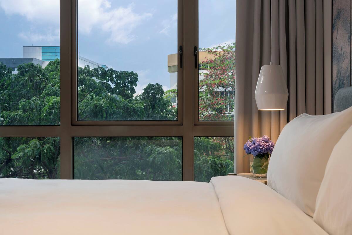 A hotel room with a large window, a bed, a bedside table with a vase of flowers, and a wall-mounted lamp.