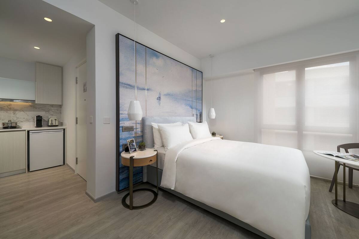 A modern hotel room with a large bed, a small kitchen area, and a dining table.